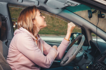 Portrait of young angry blonde woman is sitting behind the wheel of a right-handed car and shows a fist to the windshield. Side view, from inside the car. Traffic jam on the road