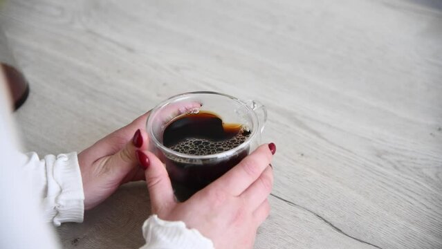 Close-up view of woman turning and holding heart shaped double wall glass mug with fresh hot black filter coffee standing on wooden table. Real time video. Coffee culture theme.