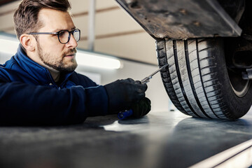 A car mechanic's shop worker checking on car tire in garage. - obrazy, fototapety, plakaty