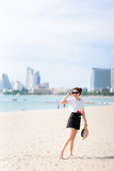 Teenage girl standing on beach looking at beautiful scenery, holding straw hat and sunglasses, smiling brightly. During summer or spring. On comfortable travel day. Empty space. Vertical image.