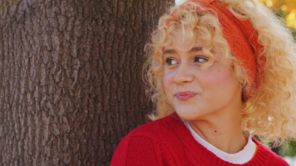 Closeup of beautiful European girl by the tree in park, in red sweater and headband looks away and smiles copy space. High quality photo