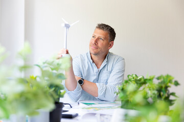 Business portrait of environmental engineer sitting at his work table holding a model of a wind turbine in his hands and is surrounded by plants