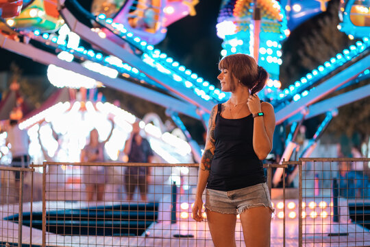 Holiday. A young happy woman with tattoos is resting in an amusement park. In the background there is a carousel glowing with neon lights