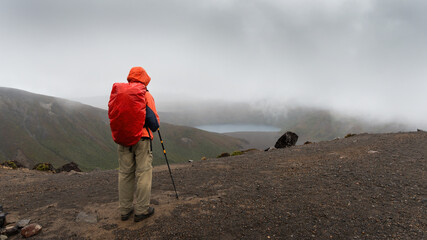 Hiking Tama Lakes track. Man looking at lower Tama lake in the mist. Tongariro National Park. - obrazy, fototapety, plakaty