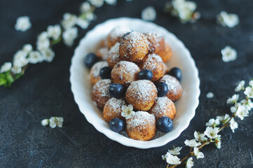 Fresh delicious homemade cottage cheese small round donuts with powdered sugar blueberries on a dark background with blossoming branches of a cherry tree. Selective focus at shallow depth of field
