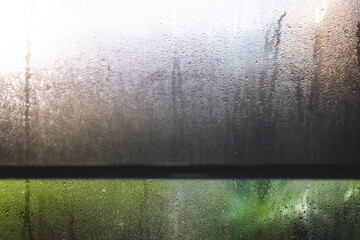 water droplets of humidity condensation on window glass seen with roller blind and backyard bokeh in the background