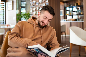 Handsome bearded hipster man relaxing holding and reading book while sitting on chair in cafe