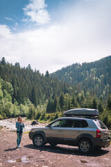 Fototapeta na wymiar Woman with car relaxing on summer road trip adventure travel. Nature travel concept with mountain background in the Ukraine. Young driver. Vertical photo. Roof rack