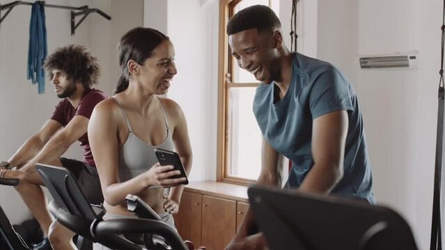 Biracial female showing black male on exercise bike funny image on cellphone at an indoor fitness gym