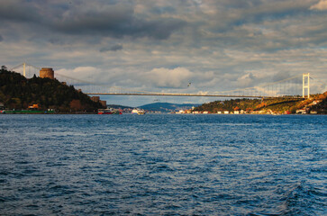Fototapeta premium Picturesque landscape view of Bosphorus strait during sunset. Cityscape with ancient towers of Rumelian Castle on green hill and Fatih Sultan Mehmet Bridge. Colorful vibrant sky. Istanbul, Turkey
