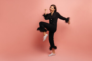 Full length young caucasian woman clenches fist, bends knee, rejoices in victory on pink background. Brunette wears black silk pajama shirt and pants. Happy morning concept