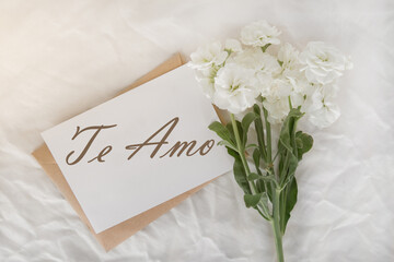 a white small bouquet on a white sheet, a craft envelope and a card with the inscription I love you in Spanish - te amo