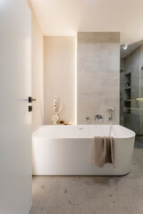 Modern bright bathroom with lamella wall. Big white bath with silver faucet and brown towel.