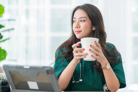 relax after work ,asian female doctor hand hold hot coffee drink cup casual relax after working hard telemedicine in clinic while wear uniform