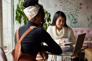 Two multicultural female students work on a project on a laptop while sitting in an internet cafe.