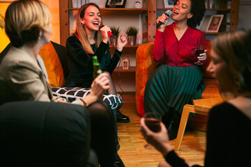 A multicultural group of female friends sings karaoke while drinking alcohol in the living room.