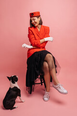 Cute young girl teaches commands to her small dog while sitting in studio on pink background. Brunette with bob haircut wears red jacket and black skirt. Lifestyle concept, pets