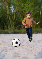 Funny Little Boy Playing with Black and White Soccer Ball in Park on Sand. Kid Running Up to Kick Ball With His Foot