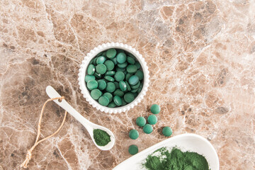 Fototapeta na wymiar Organic green spirulina powder and green algae tablets in a white bowl and spoon on a marble table. top view. food source of vitamin protein.
