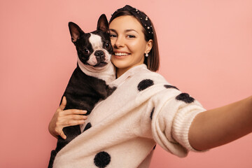 Young caucasian brunette hugging boston terrier dog making photo on pink background. Owner and pet have affection for each other. Animal love concept