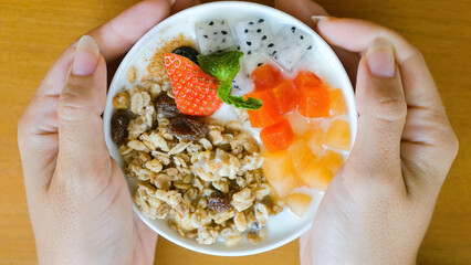 Woman hands hold bowl of homemade granola with yogurt, raisins, fresh berries, strawberry, diced melon and papaya on wooden table. Healthy breakfast food, clean eating, dieting concept. Top view.