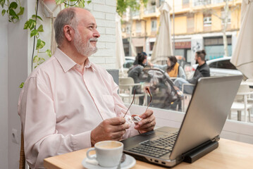 an older smiling man inside a coffee shop looking out the window, holding his eyeglasses while resting from working on his laptop