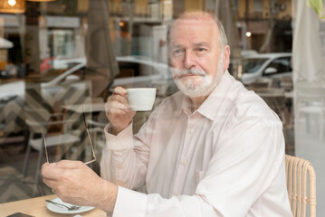 an older man sitting inside a coffee bar looking out the window holding a cup of coffee and his eyeglasses