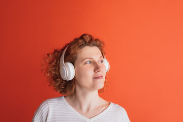 Portrait of calmly looking red-haired woman listening to music in white headphone isolated over red studio background