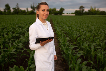 Young woman agronomist in white coat checks growth of corn in field. Farmer takes notes on tablet....