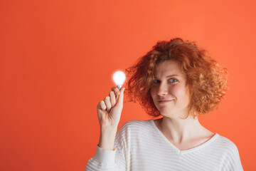 Portrait of young red-haired woman rising lightbulb, having creative idea isolated over red studio background