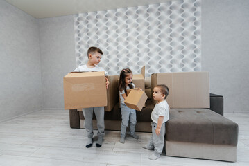Fototapeta na wymiar Brothers and sister are playing and enjoying moving to a new apartment against the background of cardboard boxes and a sofa. Purchase of real estate. Housewarming, delivery.