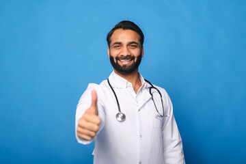 Cheerful Indian male doctor with a stethoscope in a medical gown posing isolated over blue background, showing a thumbs-up. Smiling and welcoming healthcare worker looking at the camera