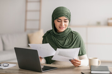 Happy young Arab woman in hijab using laptop, looking through business documents during online work meeting at home