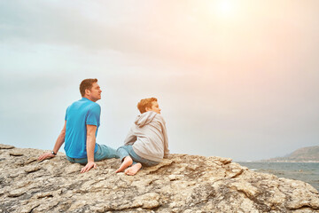 Happy family, father and son bonding, sitting on stone by the sea looking at view enjoying summer vacation. Togetherness Friendly concept
