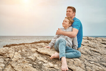 Happy family, father and son bonding, sitting on stone by the sea looking at view enjoying summer vacation. Togetherness Friendly concept