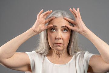 Portrait of senior woman dissatisfied with wrinkles on her forehead, touching face on grey studio background