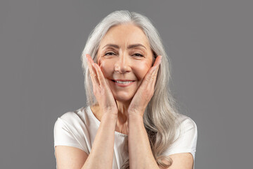 Portrait of charming senior woman looking at camera and smiling, touching smooth skin on her face