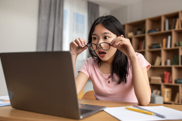 Unbeliveable. Shocked asian lady in eyeglasses looking at laptop computer screen, sitting at desk at home
