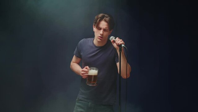Drunk man singing on stage and drinking beer. Young guy in karaoke, male person speaking with audience using microphone.