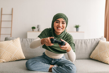 Happy young Arab woman in hijab playing online video game with joystick, sitting on couch at home, copy space © Prostock-studio