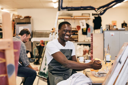 Portrait of happy young male student in apron sitting at table in art class