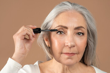 Beauty portrait of charming senior woman applying mascara on her lashes over brown studio background