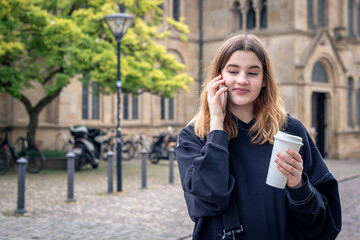 A young woman drinking coffee and talking on the phone on a walk in the city.