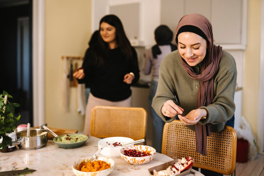 Smiling young woman in hijab garnishing food with pomegranate in kitchen at home