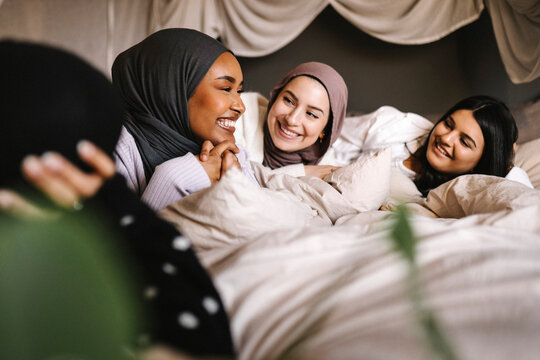 Happy young women in hijabs looking at smiling female friend on bed at home