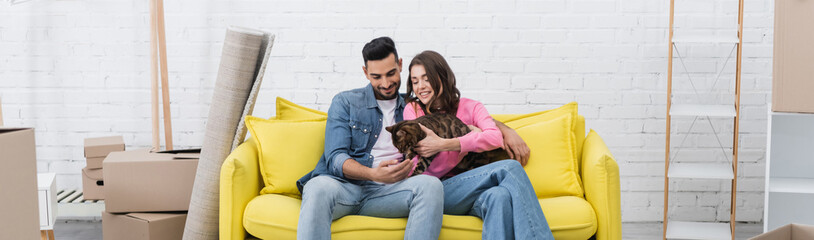 Cheerful interracial couple petting bengal cat on couch near packages at home, banner.
