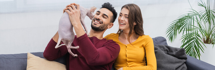 Cheerful muslim man holding sphynx cat near girlfriend on couch, banner.