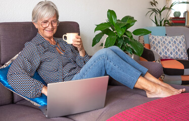 Relaxed elderly woman working on laptop at home sitting on sofa in living room. Senior lady holding a cup of coffee enjoying tech and social