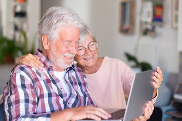 Joyful beautiful senior couple smiling at home browsing together on laptop. Active elderly Caucasian couple enjoying technology and social