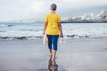 Rear view of senior woman barefoot walking on sea shore holding shoes and hat in hands.Elderly woman on a black sand beach enjoying vacation and freedom